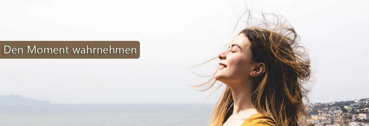 Frau steht am Meer und der Wind bläst ihr durch die Haare. Beschriftung: Den Moment wahrnehmen.